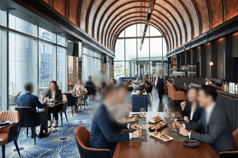 Patrons dressed in business attire dine in a sophisticated, modern restaurant with high, arched wooden ceilings, expansive windows, and a vibrant blue patterned carpet, evoking the atmosphere of a beloved gathering place.