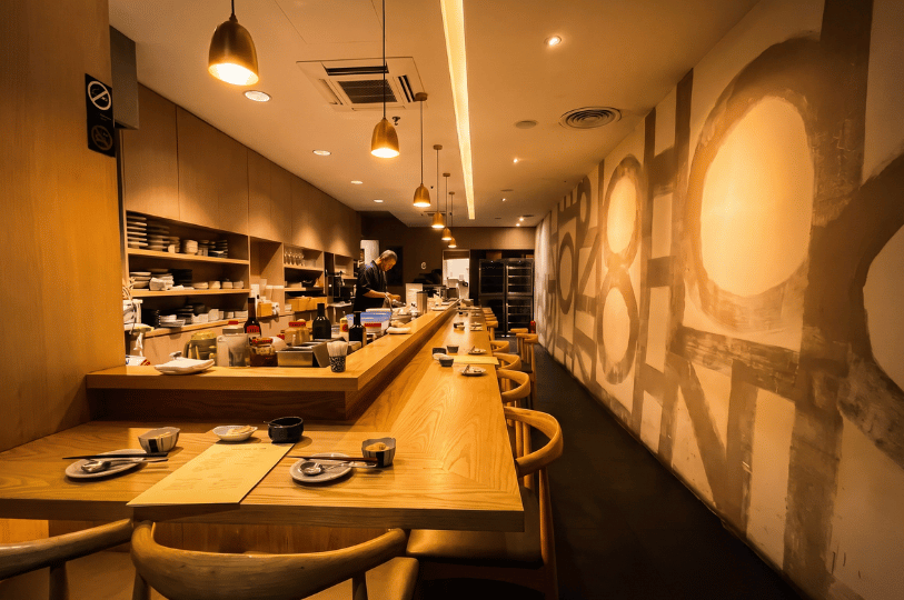 An empty, long wooden L-shaped dining counter under warm hanging lights at Bistro du Le Pin, featuring abstract wall art and a chef preparing food in the background.