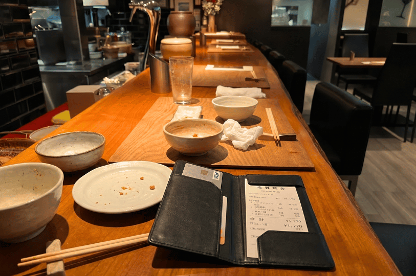 A black leather bill presenter sits on a wooden restaurant counter. The counter is scattered with empty ceramic bowls, a used plate with crumbs, wooden chopsticks, and crumpled napkins from a finished meal. The background shows a warmly lit, blurred view of the restaurant's bar area and empty dark seating.