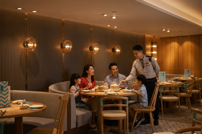 A warm, well-lit scene of a family of four enjoying a meal at a modern, upscale Chinese restaurant. They are seated at a round wooden table in a cozy curved booth. A smiling waiter in uniform is serving them, lifting the lid of a steaming bamboo dim sum basket. The restaurant features elegant wood-paneled walls with decorative glass sconces, capturing the joy of shared family dining.