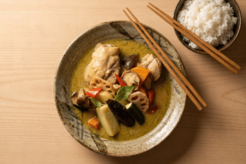 Top-down view of a rustic ceramic bowl filled with vibrant Hokkaido soup curry. The rich, green spiced broth features tender pieces of chicken, sliced lotus root, mushrooms, zucchini, eggplant, carrots, and bell peppers, topped with fresh basil. Wooden chopsticks rest across the bowl, placed next to a small side dish of fluffy white rice on a light wooden table.