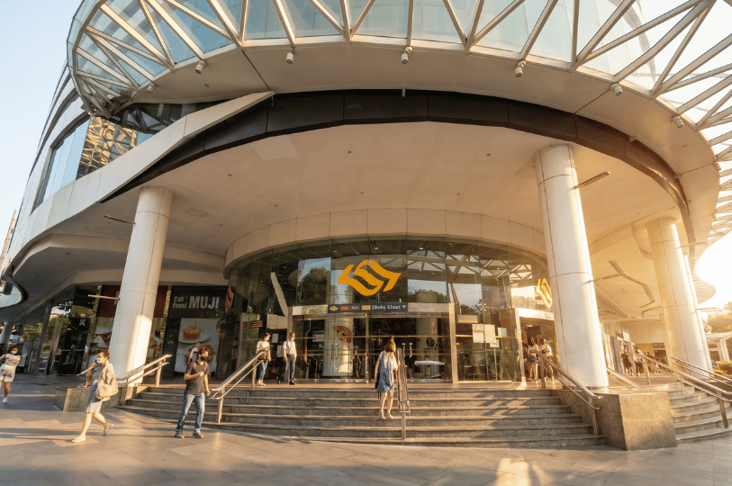 Exterior view of the Dhoby Ghaut MRT station entrance under a large, modern glass canopy with pedestrians walking by in the sunlight.