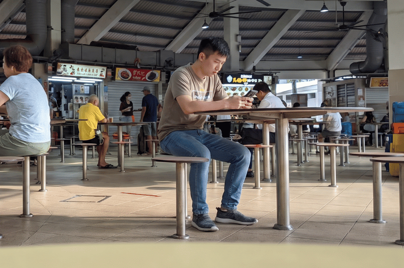 A young man sitting alone on a fixed stool at a round table in a spacious Singaporean hawker centre, quietly engrossed in his smartphone. The candid shot captures an everyday urban moment, showing the food court's tiled floors, high ceilings with large industrial fans, and the lively background of brightly lit food stalls and diverse patrons sharing the communal dining space.