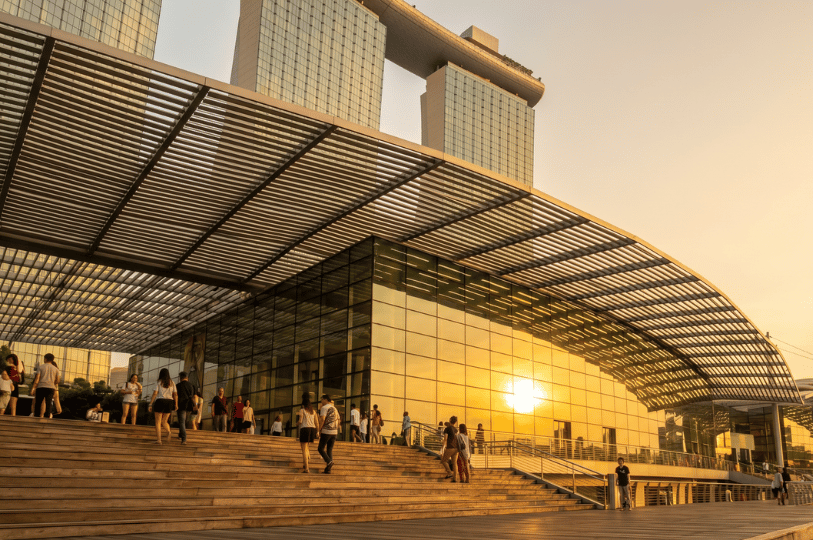 A golden sunset reflects brilliantly off the glass facade of a modern pavilion. In the foreground, people walk along wide wooden steps beneath a large, slatted canopy, while the iconic, ship-topped towers of the Marina Bay Sands resort rise into the evening sky in the background.