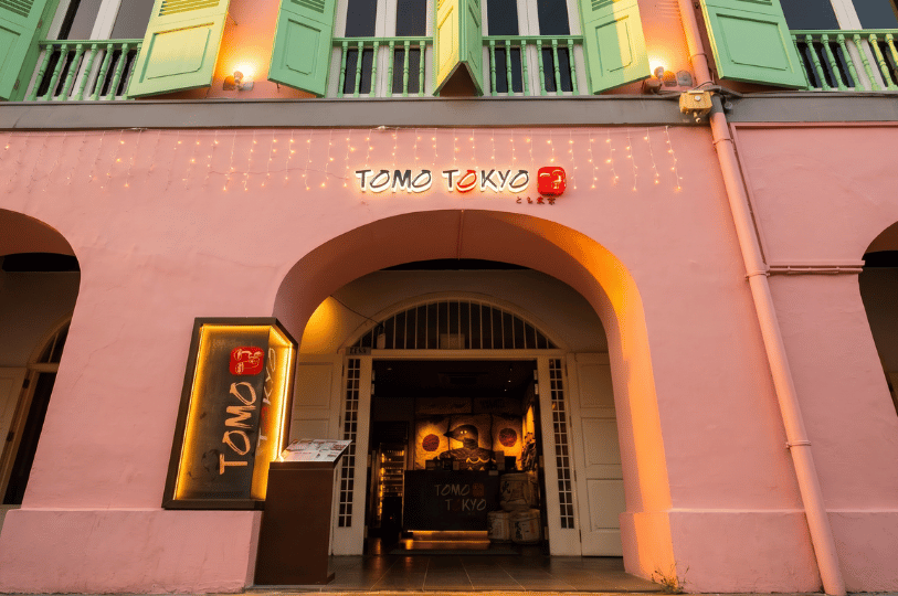 Exterior storefront of Tomo Tokyo at Clarke Quay, featuring a bright pink traditional shophouse facade with large arched entryways, light green shutters, and a glowing illuminated restaurant sign.