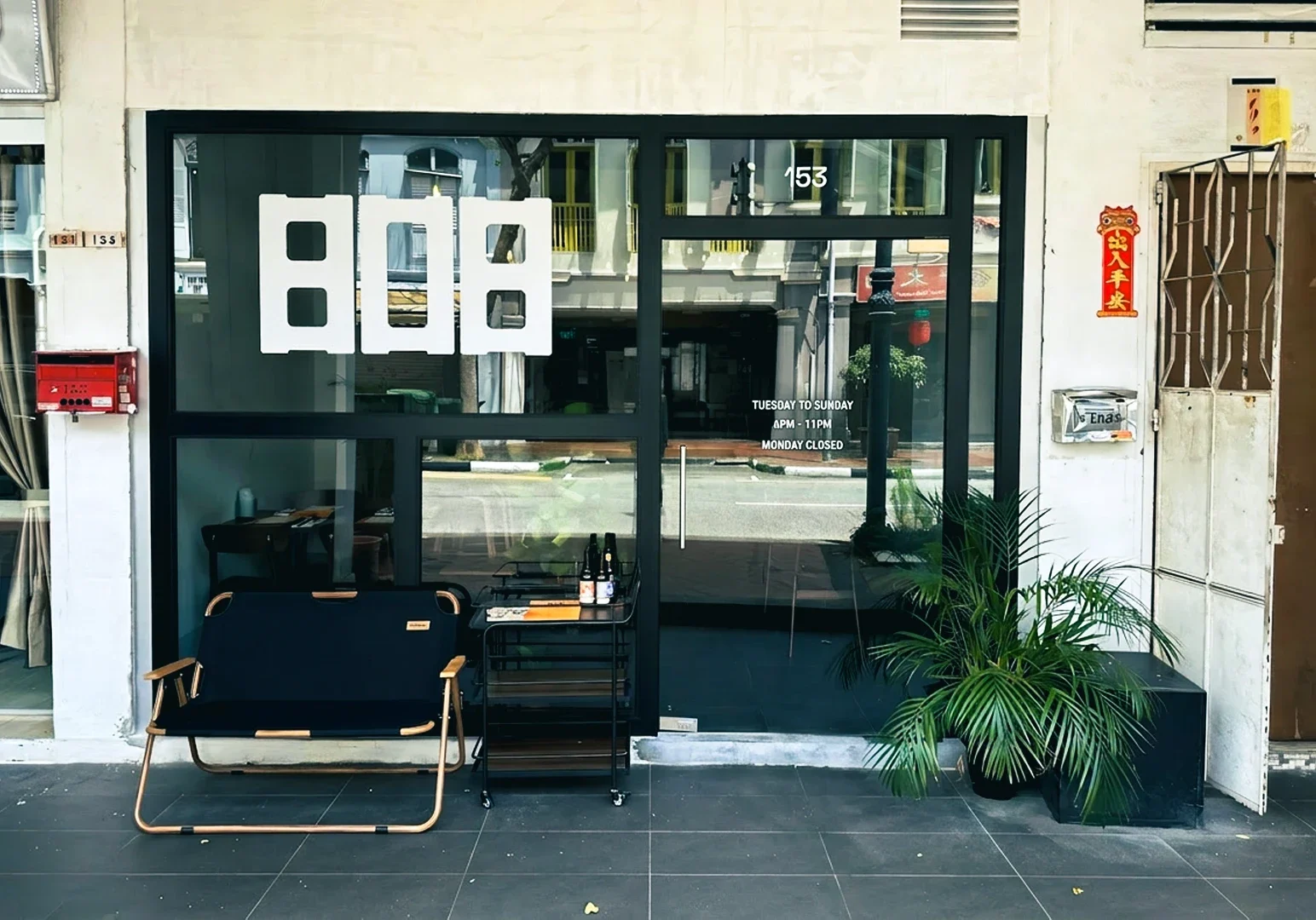 Front entrance of 808 Eating House with large glass windows, minimalist signage, a small bench, potted plants, and a side table near the doorway.