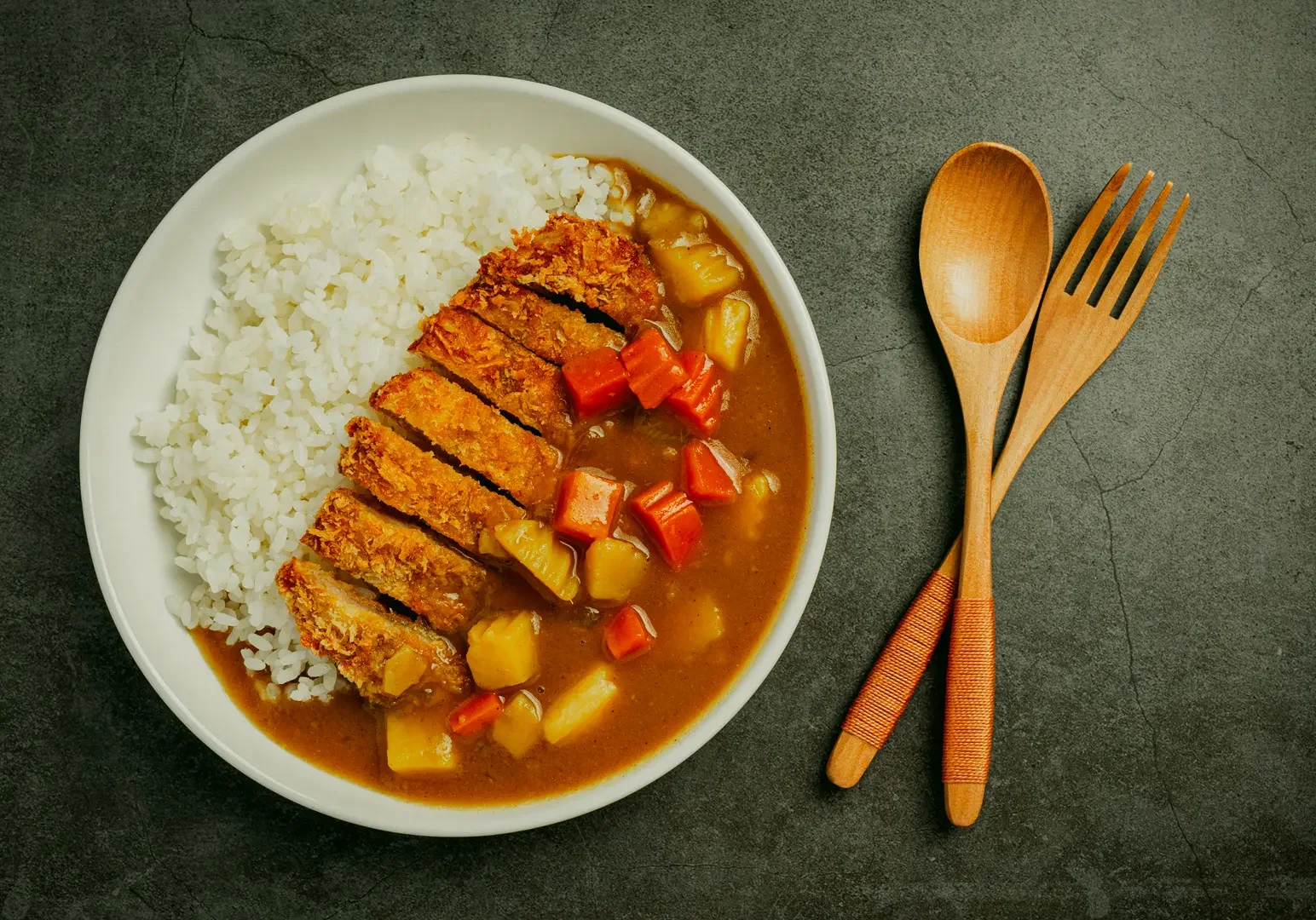 Top-down, flat-lay shot of a Japanese pork katsu curry rice dish served in a white bowl on a dark stone-textured surface, featuring neatly arranged white rice beside thick brown Japanese curry sauce with visible diced carrots and potatoes, topped with sliced golden-brown breaded pork cutlet. A wooden spoon and wooden fork rest parallel on the right side of the frame, with soft, even lighting highlighting the glossy curry texture and crisp panko coating, creating a clean, editorial food presentation.