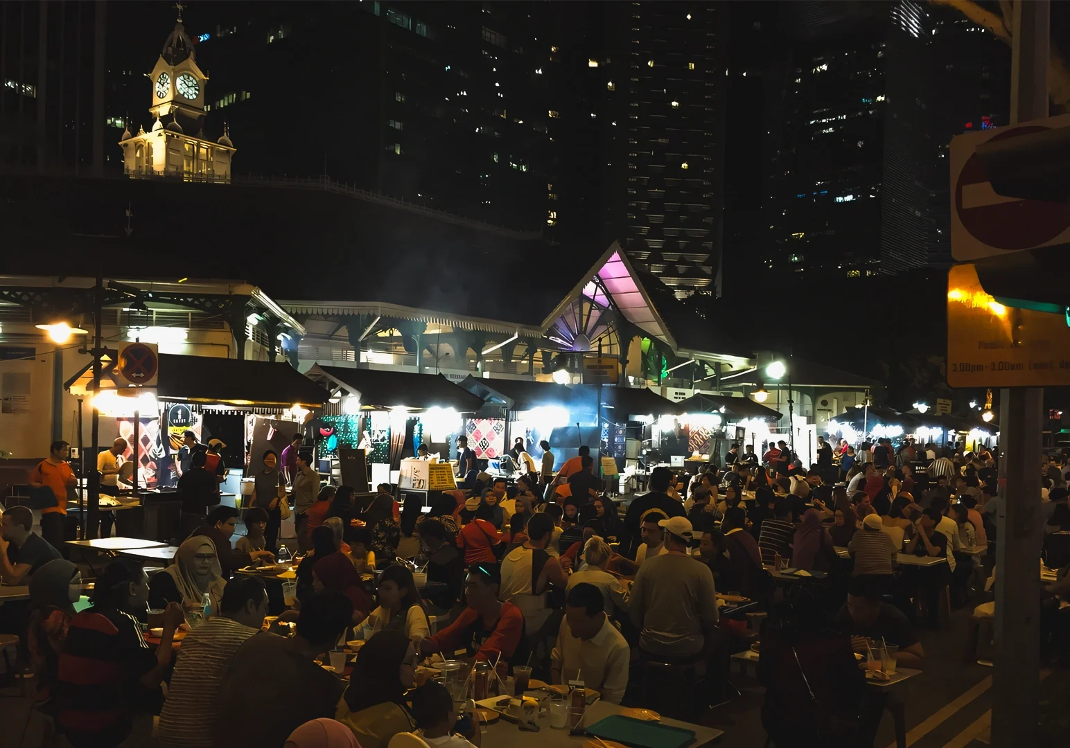 1 Chasing After-Hours Bites Wide‑angle night shot of a bustling street food market filled with outdoor dining tables, glowing food stalls, and city skyscrapers in the background, capturing vibrant urban nightlife and after‑hours dining culture.