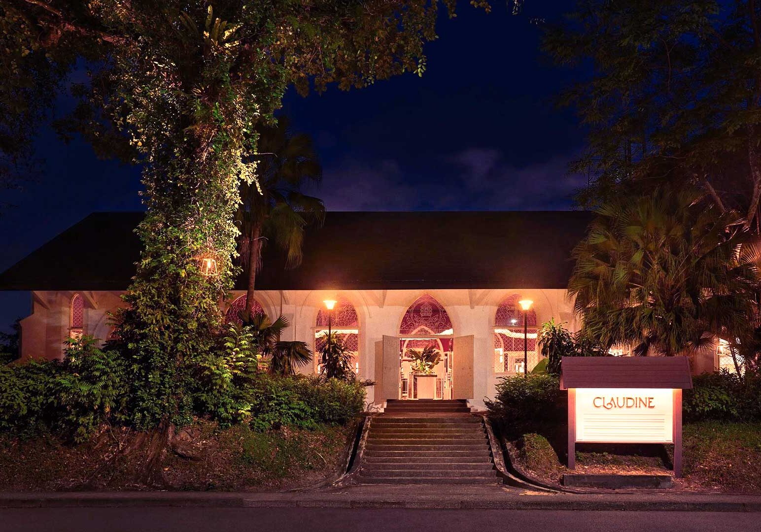 Exterior of a restaurant at night featuring a steep pitched roof, illuminated arched windows with red patterns, and a staircase framed by plants and trees.