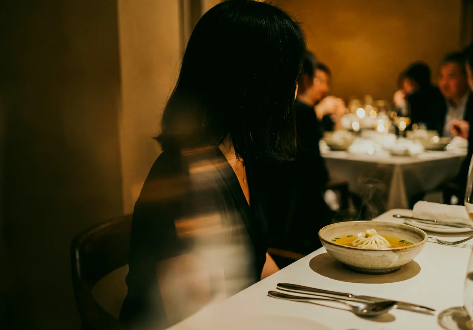 Eye-level shot of a refined fine dining restaurant scene with a diner seated beside a bowl of plated comfort food, warm ambient lighting and elegant table setting highlighting modern upscale cuisine.