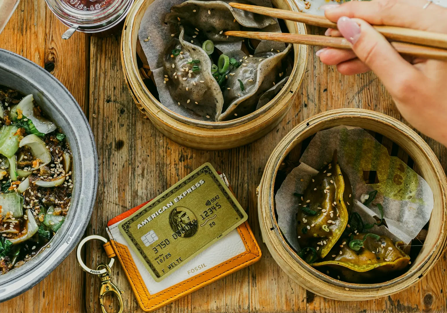 Top-down overhead shot of shared Asian dishes including dumplings on a wooden table, with chopsticks and payment cards, representing budget-friendly dining and meal deals.
