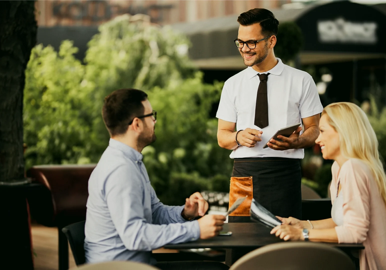 1 Dining Elegance Without the Stiffness Eye-level medium shot of a server standing beside a café table while presenting a menu tablet to seated diners in an outdoor dining area surrounded by greenery, illustrating relaxed yet refined restaurant service.