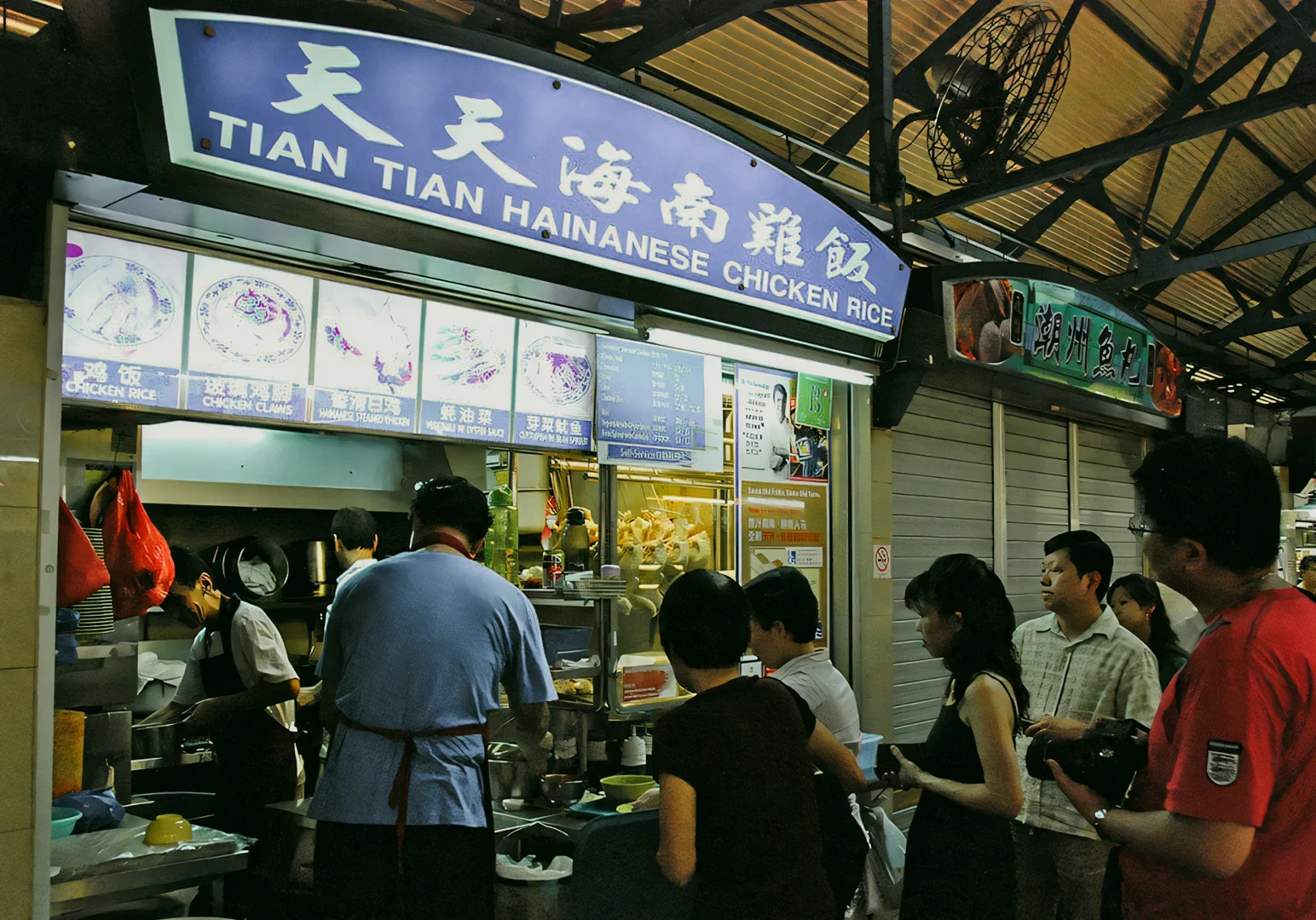 Eye-level wide-angle shot of Tian Tian Hainanese Chicken Rice hawker stall, showing customers queueing at a brightly lit food counter inside a covered market.