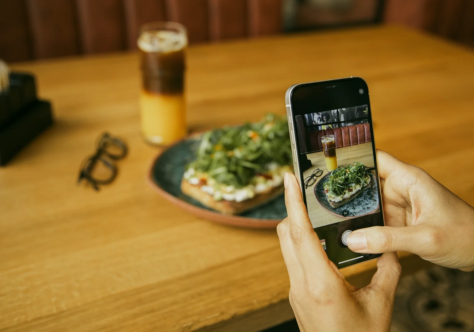 Over‑the‑shoulder close‑up shot of a person photographing a plated dish and coffee on a wooden restaurant table using a smartphone, highlighting dining as a shared food experience.