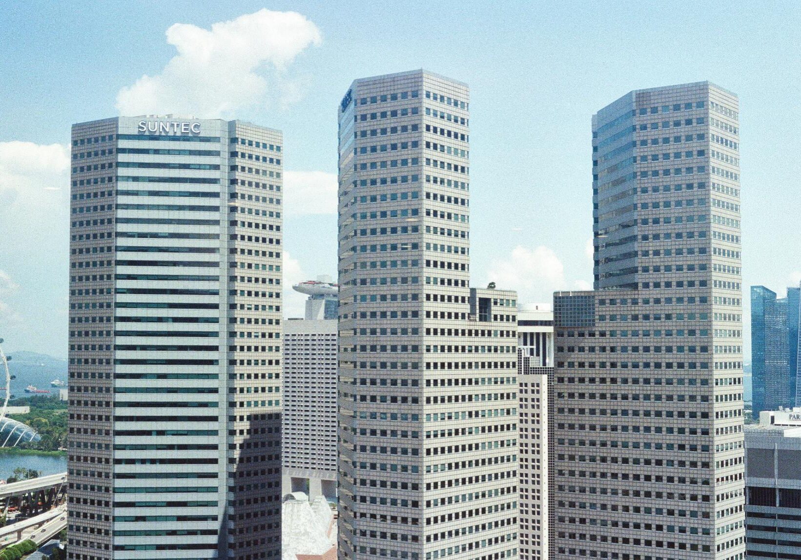 This image captures the distinctive high-rise office towers of Suntec City in Singapore, characterized by their geometric, faceted architecture and repetitive window patterns. In the background, other iconic landmarks like the Marina Bay Sands and the Singapore Flyer are visible under a bright, clear sky.