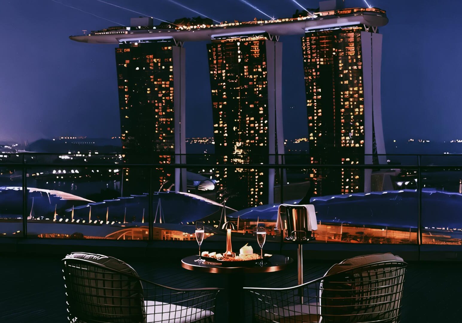Romantic rooftop dining setup overlooking Marina Bay Sands at night, featuring candlelit wine glasses, a dessert platter, and panoramic views of Singapore’s illuminated skyline.