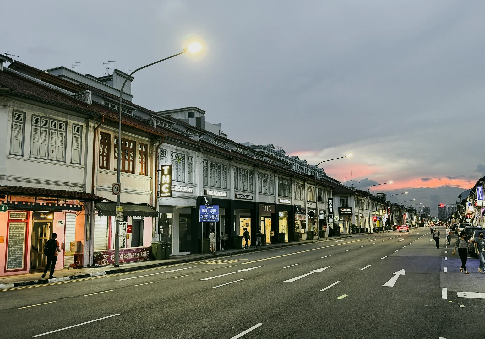 Heritage shophouses along Geylang Singapore at dusk with glowing streetlights and quiet road, shot at a low-angle leading-line perspective.