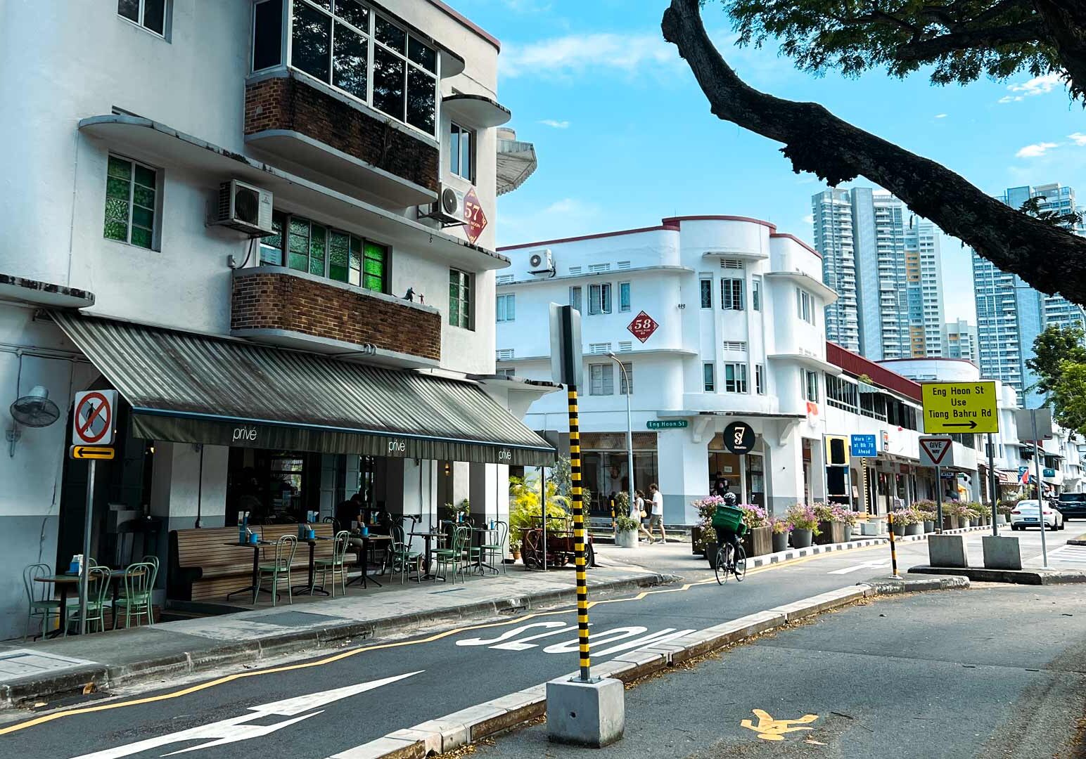 Quiet urban street lined with low-rise white Art Deco–style buildings, outdoor cafés, bicycle lanes, and high-rise skyscrapers in the distance.