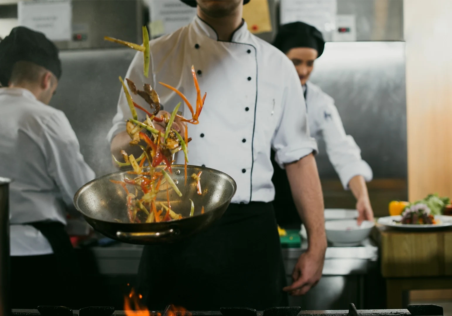 Medium shot at eye level of a professional chef tossing vegetables in a hot pan over an open flame inside a commercial restaurant kitchen.