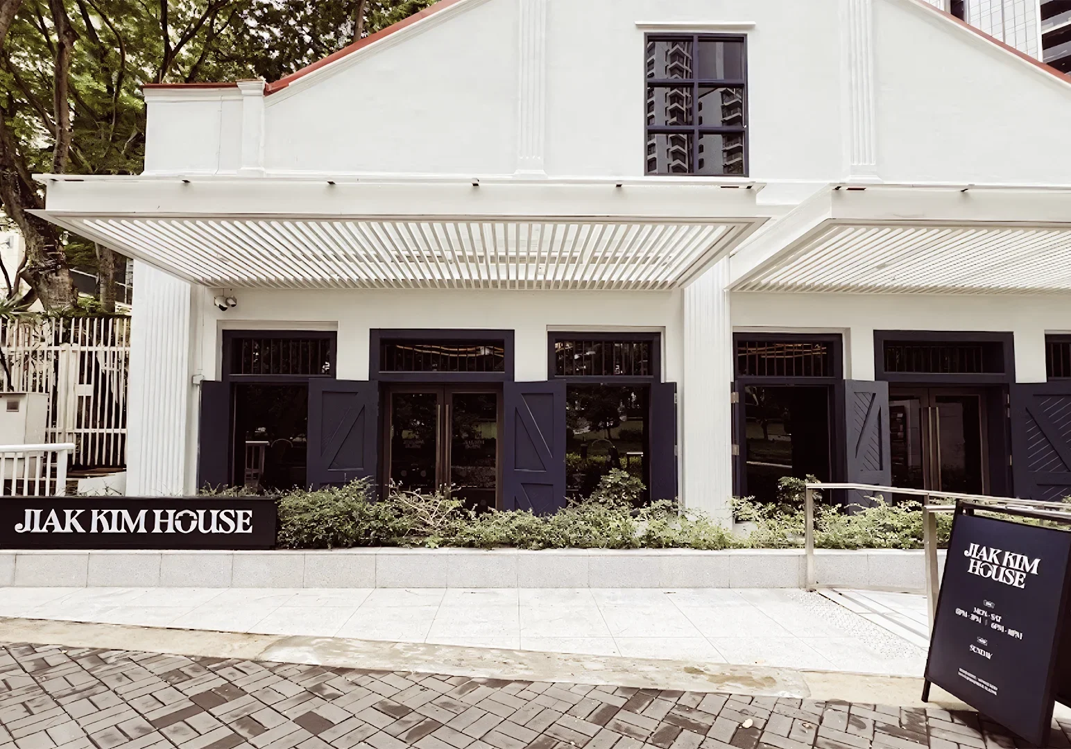 1 Jiak Kim House Singapore Front facade of Jiak Kim House with white colonial-style architecture, black-framed windows, and a walkway leading to the entrance surrounded by greenery.