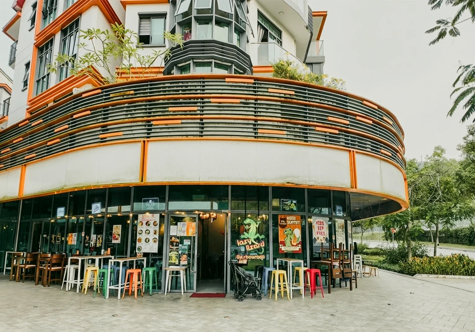 1 Lazy Lizard Flora Vista Wide-angle eye-level shot of a colorful corner restaurant exterior with curved balcony railings, glass storefront, and outdoor seating in a modern urban setting.
