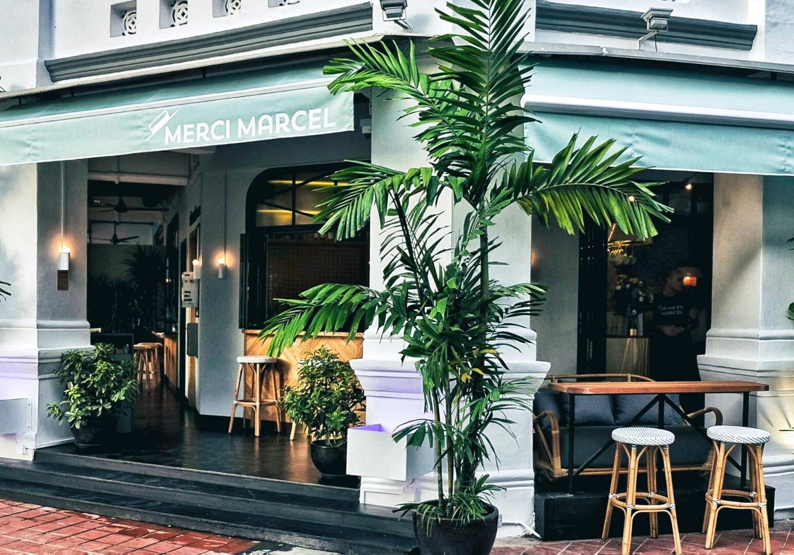 Exterior of Merci Marcel café with teal awnings, large potted plants, and outdoor seating in front of a white colonial-style building.