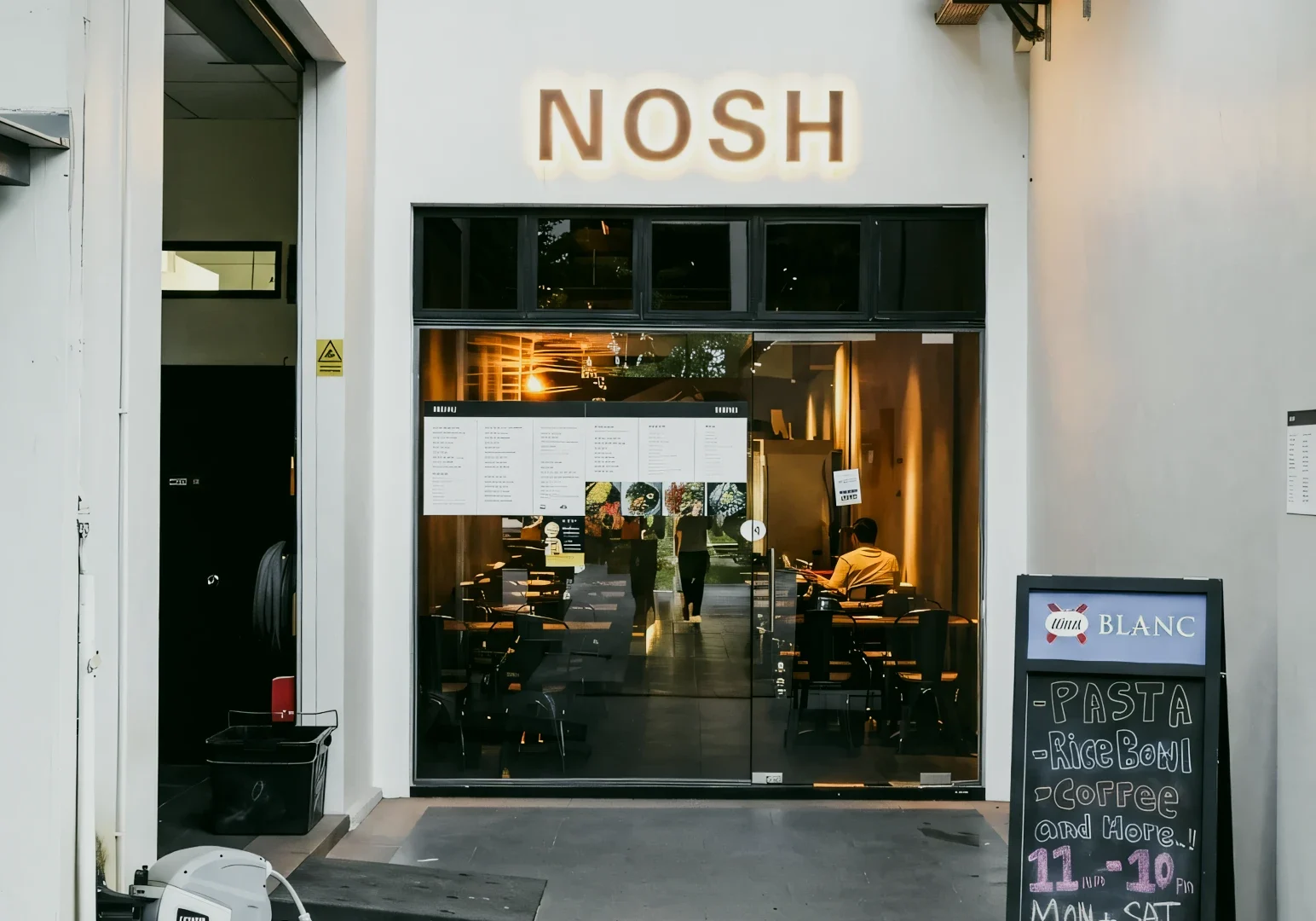 Eye-level straight-on shot of the NOSH restaurant storefront with glass entrance, illuminated signage, visible indoor seating, and a sidewalk menu board.