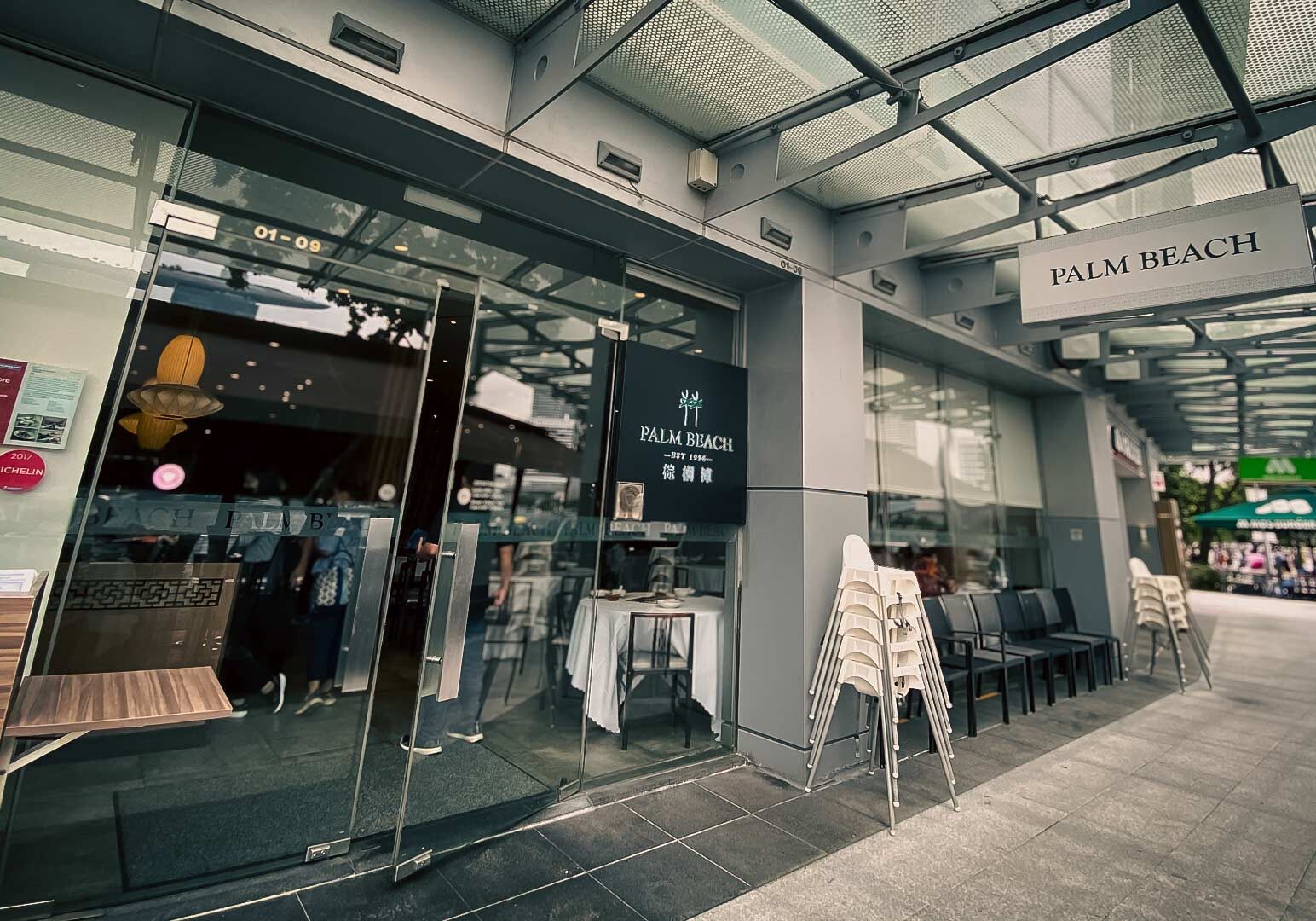 1-Palm-Beach-Seafood Entrance of Palm Beach Seafood restaurant featuring a modern glass storefront, signage above the doorway, and outdoor seating stacked alongside the entrance.