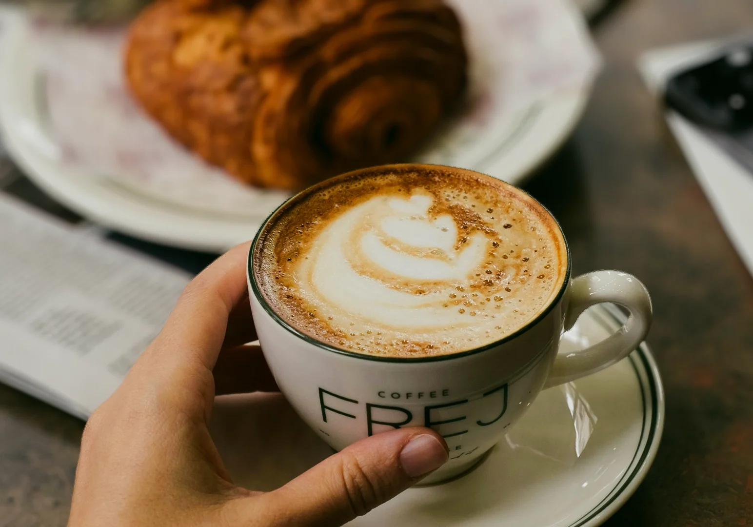 1 Restaurant Hopping Close‑up eye‑level shot of a hand holding a cappuccino with latte art in a ceramic cup, paired with a flaky croissant on a café table, showcasing specialty coffee culture, café hopping, and relaxed brunch moments.