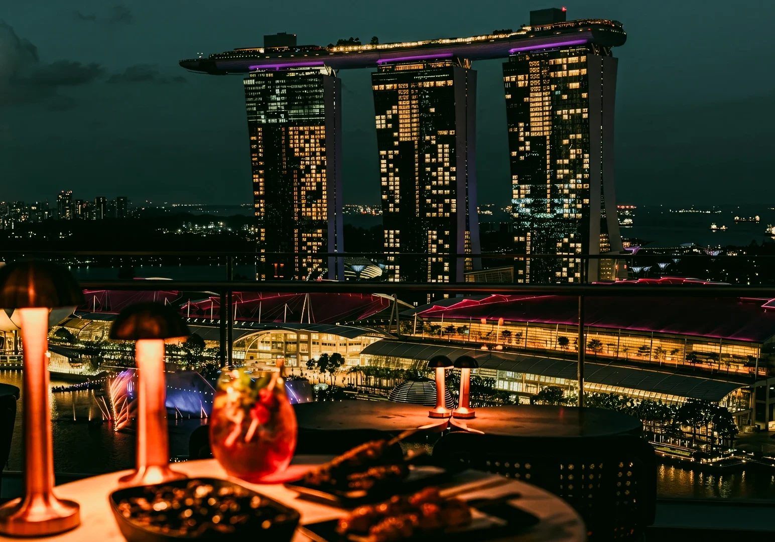 Eye‑level wide‑angle interior shot of a luxury restaurant dining table beside floor‑to‑ceiling windows overlooking Marina Bay Sands and Singapore harbor.