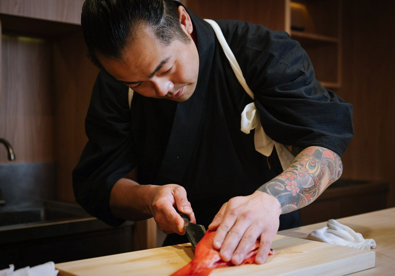 Japanese chef filleting red fish on wooden board, tattoo sleeve visible, traditional black robe with white sash.