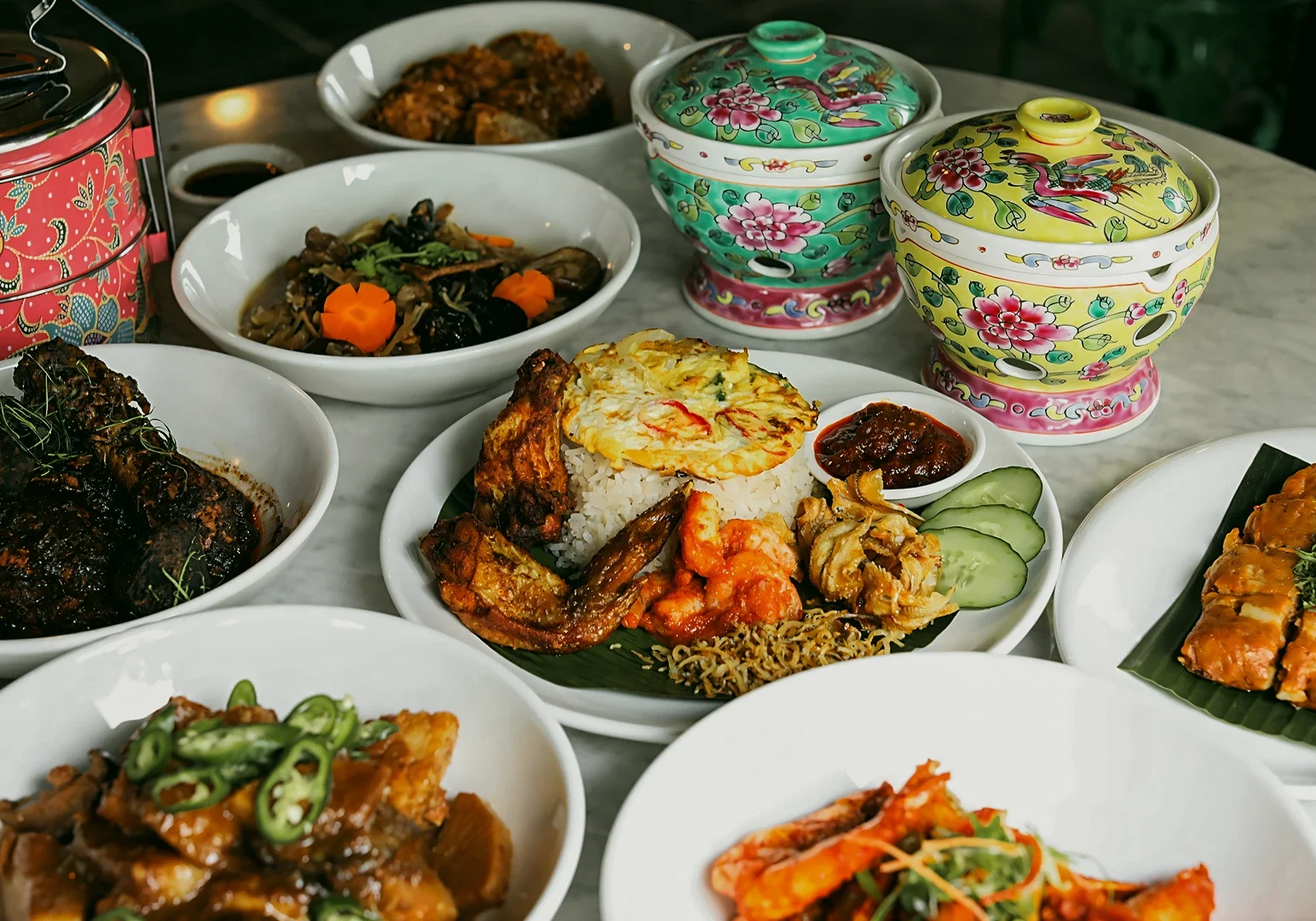 1 The Dedication Behind Peranakan Cuisine Singapore Through a Chef’s Work Eye-level slightly overhead shot of a Peranakan cuisine spread in Singapore featuring Nyonya dishes, rice with sambal, prawns, braised meats, and traditional ceramic serving ware on a marble table under warm lighting.