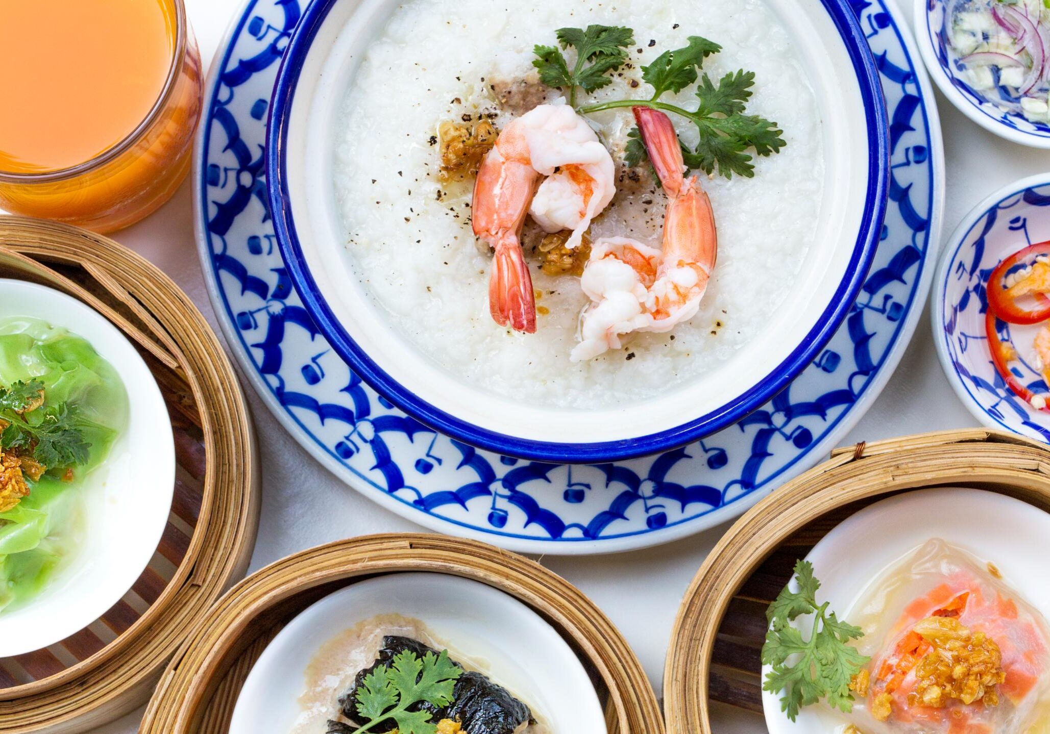 This image features a top-down view of an oriental breakfast set, centered around a bowl of shrimp congee garnished with cilantro and black pepper. The meal is complemented by several bamboo steamer baskets containing various dim sum, along with small side dishes of chilies, aromatics, and a glass of orange juice.