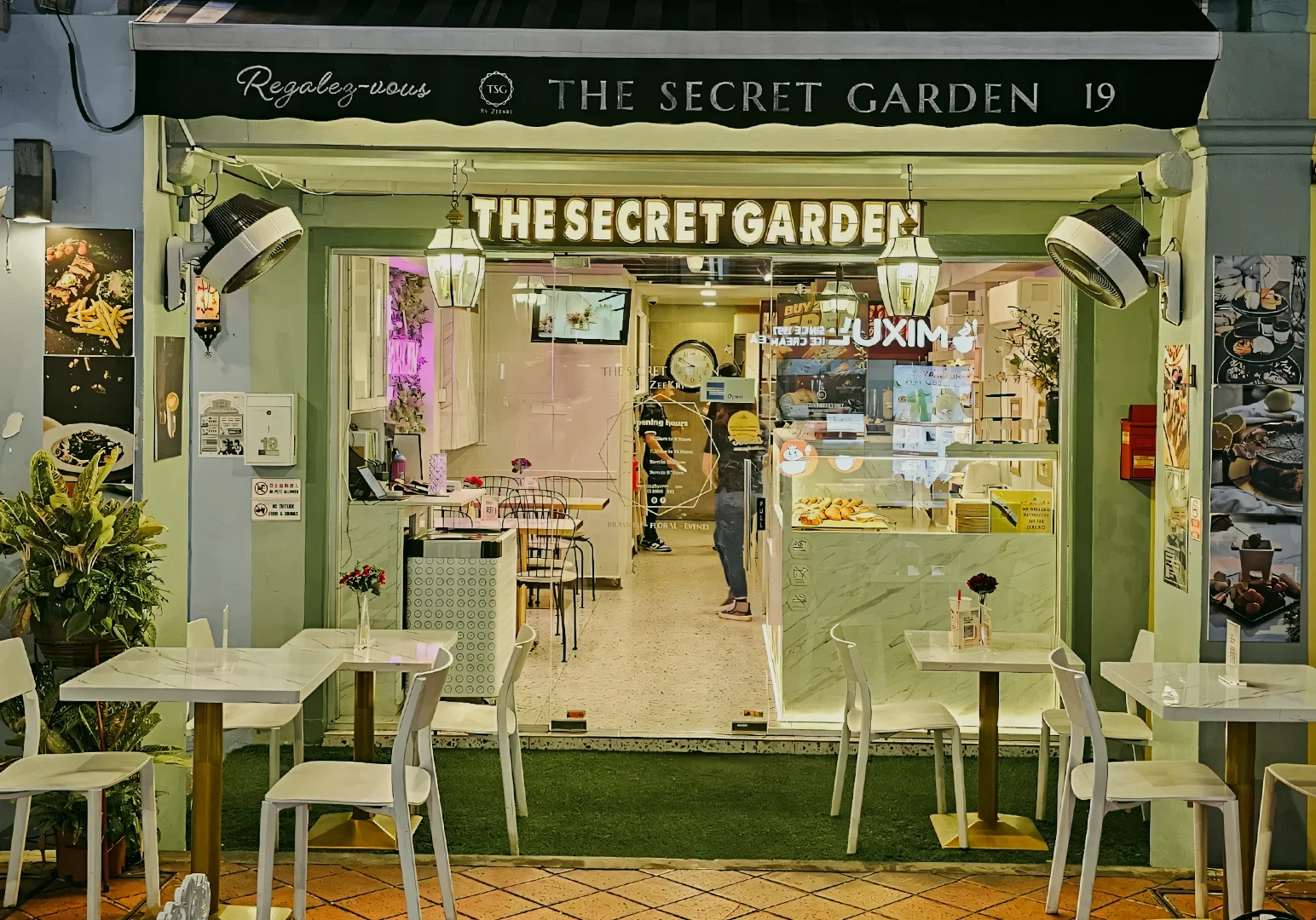 1 The Secret Garden By Zeekri Straight-on street-level shot of “The Secret Garden” café storefront with green façade, outdoor white tables and chairs, warm interior lighting, and clear signage, presenting an inviting boutique café exterior at night.