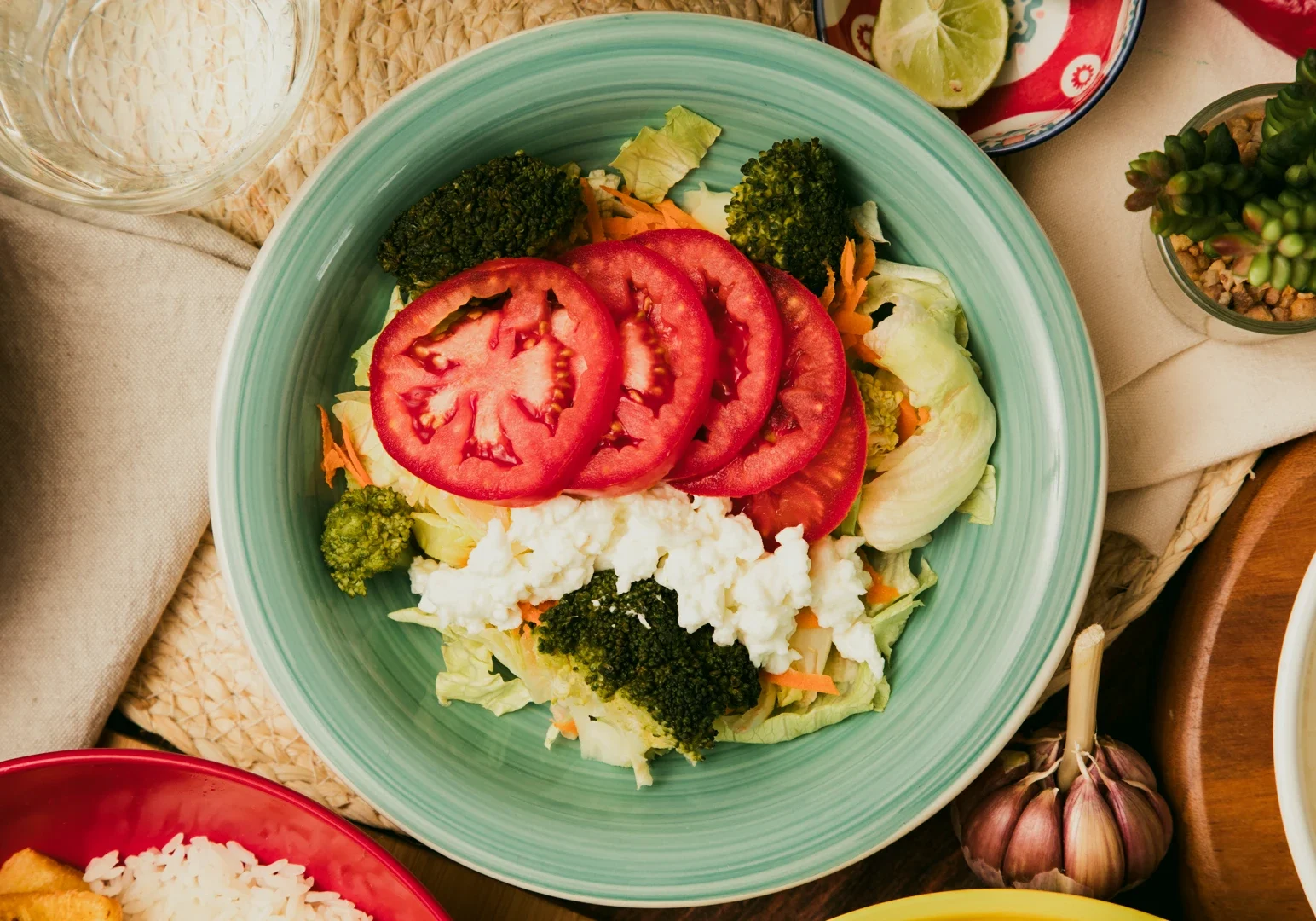 Overhead top‑down close‑up shot of a vegetarian salad bowl with sliced tomatoes, broccoli, lettuce, rice, and mixed vegetables arranged on a light blue plate on a rustic table setting.