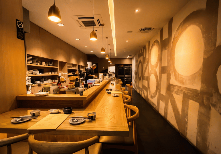 An empty, long wooden L-shaped dining counter under warm hanging lights at Bistro du Le Pin, featuring abstract wall art and a chef preparing food in the background.