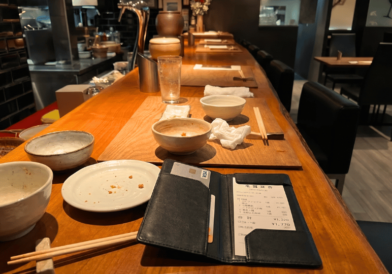 A black leather bill presenter sits on a wooden restaurant counter. The counter is scattered with empty ceramic bowls, a used plate with crumbs, wooden chopsticks, and crumpled napkins from a finished meal. The background shows a warmly lit, blurred view of the restaurant's bar area and empty dark seating.