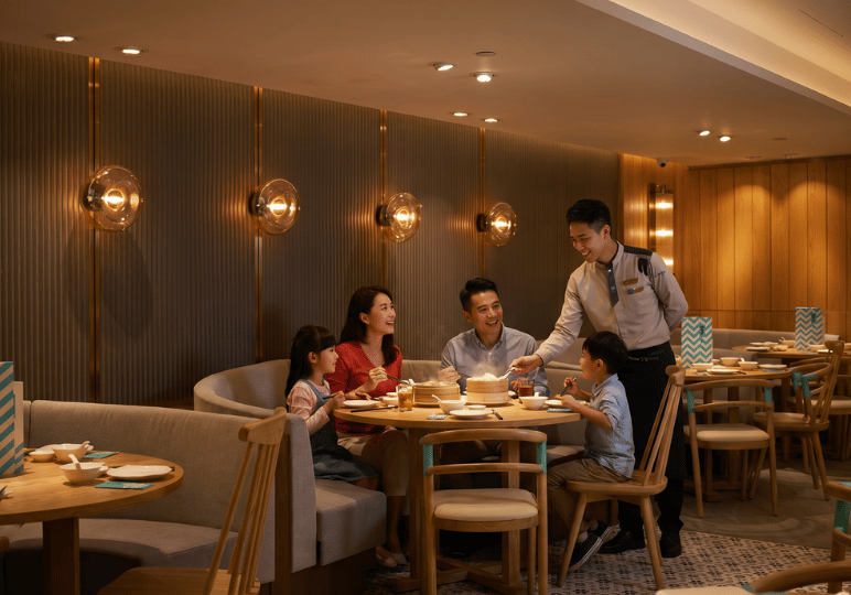 1-family-dining A warm, well-lit scene of a family of four enjoying a meal at a modern, upscale Chinese restaurant. They are seated at a round wooden table in a cozy curved booth. A smiling waiter in uniform is serving them, lifting the lid of a steaming bamboo dim sum basket. The restaurant features elegant wood-paneled walls with decorative glass sconces, capturing the joy of shared family dining.