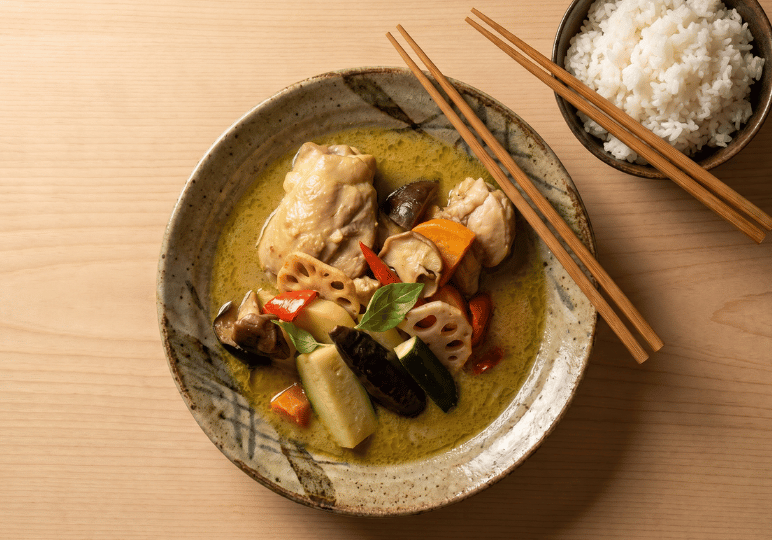 1-finding-stillness-in-a-bowl-of-soup-curry Top-down view of a rustic ceramic bowl filled with vibrant Hokkaido soup curry. The rich, green spiced broth features tender pieces of chicken, sliced lotus root, mushrooms, zucchini, eggplant, carrots, and bell peppers, topped with fresh basil. Wooden chopsticks rest across the bowl, placed next to a small side dish of fluffy white rice on a light wooden table.