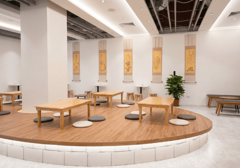 A serene, minimalist cafe interior at Hvala Singapore featuring low wooden tables and round floor cushions on a curved, raised wooden platform, with traditional Japanese hanging scrolls decorating the white walls.