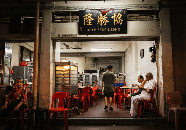 A nighttime view of the traditional Heap Seng Leong kopitiam illuminated by pale interior fluorescent lights. Patrons unwind in bright red plastic chairs, including an older woman resting by the entrance and two men quietly checking their phones, capturing a subdued and restful evening atmosphere.