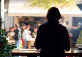 1-li jun-Writers Silhouette of a person sitting at a wooden cafe table, looking out at a blurred, busy outdoor market.