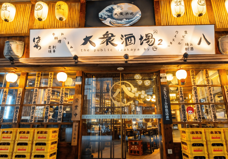 1-restaurant-interior A warm, inviting wooden storefront of a Japanese izakaya, featuring glowing paper lanterns, traditional signage, stacked yellow crates, and glass doors that reveal a cozy, softly lit interior.