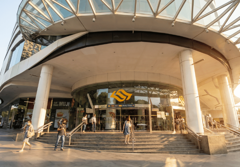 Exterior view of the Dhoby Ghaut MRT station entrance under a large, modern glass canopy with pedestrians walking by in the sunlight.
