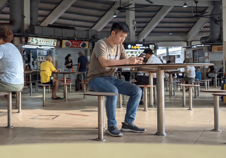 A young man sitting alone on a fixed stool at a round table in a spacious Singaporean hawker centre, quietly engrossed in his smartphone. The candid shot captures an everyday urban moment, showing the food court's tiled floors, high ceilings with large industrial fans, and the lively background of brightly lit food stalls and diverse patrons sharing the communal dining space.