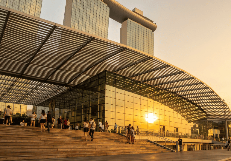 A golden sunset reflects brilliantly off the glass facade of a modern pavilion. In the foreground, people walk along wide wooden steps beneath a large, slatted canopy, while the iconic, ship-topped towers of the Marina Bay Sands resort rise into the evening sky in the background.
