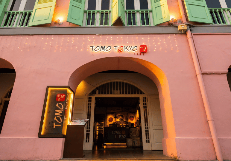 Exterior storefront of Tomo Tokyo at Clarke Quay, featuring a bright pink traditional shophouse facade with large arched entryways, light green shutters, and a glowing illuminated restaurant sign.
