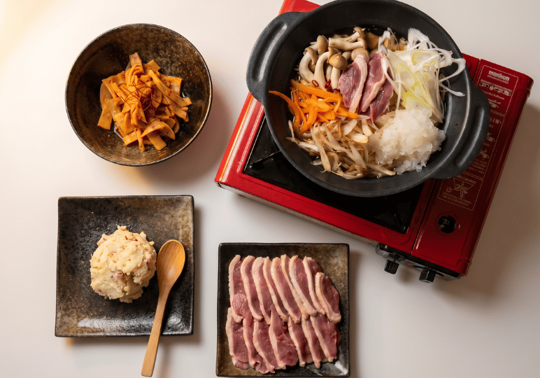 13-orchard-plaza-stalls A Japanese hot pot spread featuring a pot of sliced duck, mushrooms, and fresh vegetables on a red portable stove, accompanied by plates of raw duck, potato salad, and seasoned bamboo shoots.