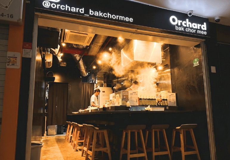 14-orchard-plaza-stalls The intimate, open-kitchen storefront of Orchard Bak Chor Mee, featuring chefs cooking amidst rising steam behind a black dining counter lined with wooden stools.