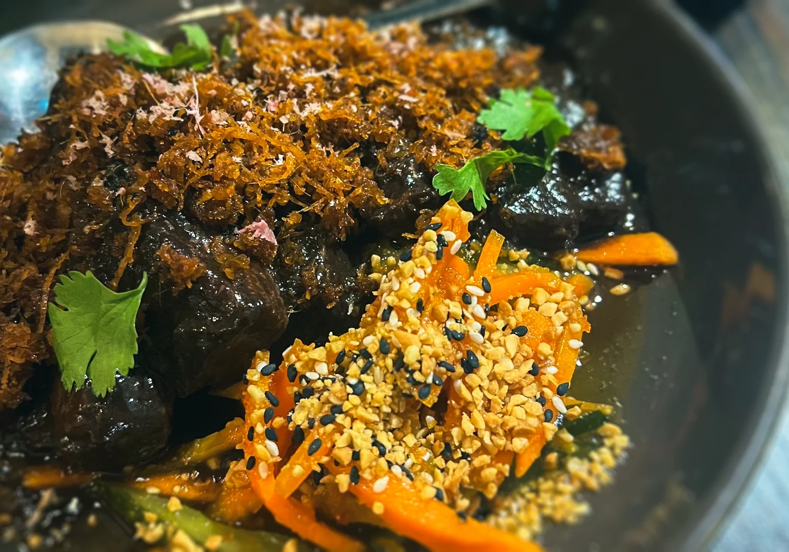 Plate of Buah Keluak pork belly featuring tender braised pork glazed in rich buah keluak sauce, topped with crispy fried shallots and cilantro, served alongside pickled carrots, cucumbers, and toasted sesame seeds.